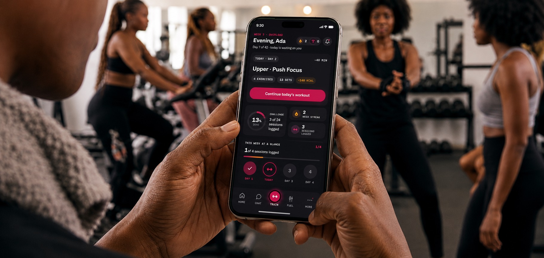 A member holding a phone showing the DP Fitness app's home screen, mid-session on a gym floor